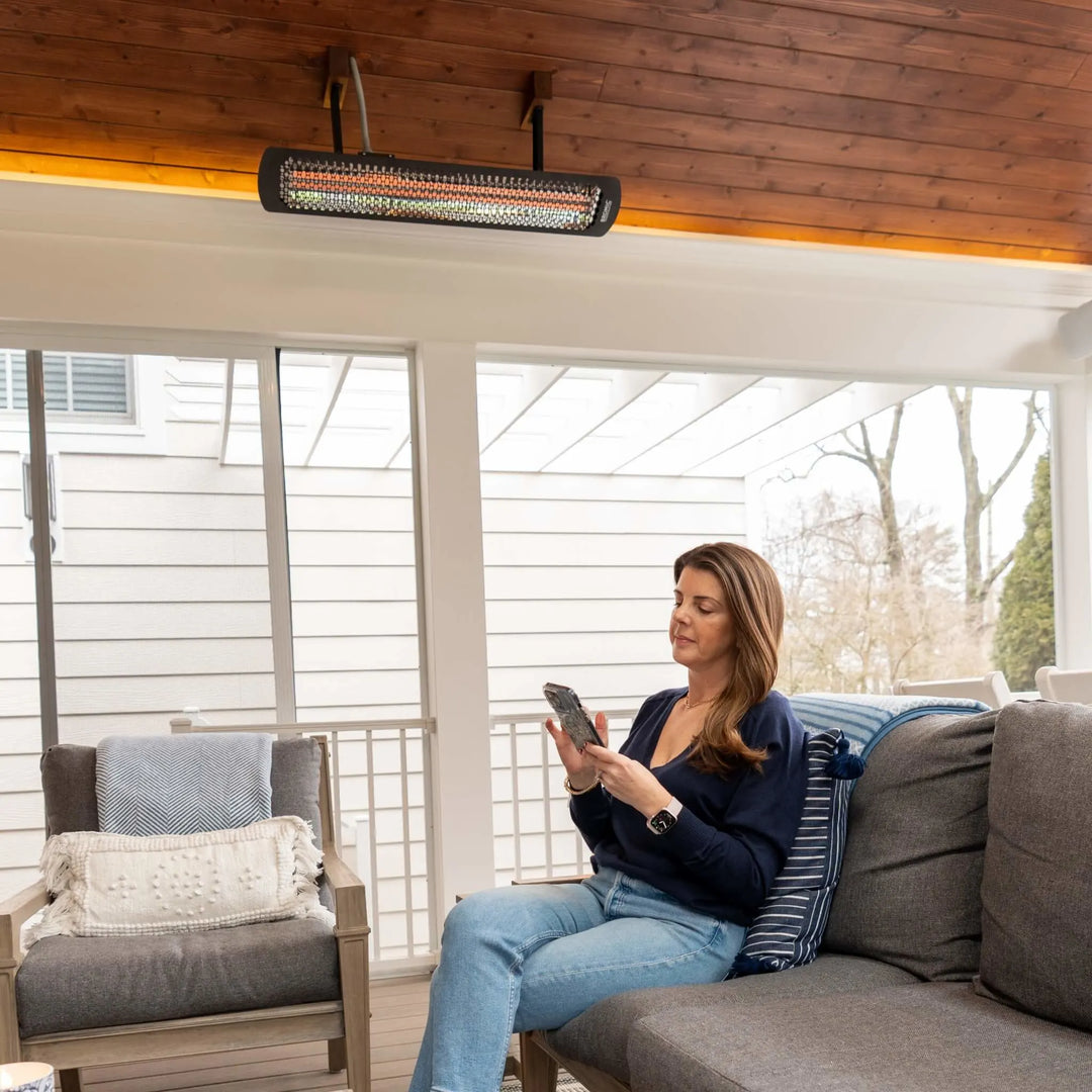 Cozy enclosed patio with a Bromic Heating Tungsten Smart-Heat Electric Heater above a comfortable seating area, where a woman relaxes and uses her smartphone beneath soft radiant warmth.