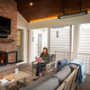 Enclosed patio space with a Bromic Heating Tungsten Smart-Heat Electric Heater installed overhead, providing warmth as a woman works on her laptop beside a lit fireplace.