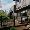Modern outdoor deck with Bromic Tungsten Electric Smart-Heat heaters mounted along a dark wood exterior wall, warming a cozy patio lounge area with sleek furniture and lush greenery.