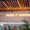 Contemporary restaurant interior featuring Bromic Tungsten Electric Smart-Heat heaters ceiling-mounted above a “MAKE IT HAPPEN” neon sign, blending warmth and industrial style for comfortable dining.