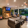 Inviting backyard patio with Bromic Tungsten Electric Smart-Heat heaters installed overhead, illuminating a wicker furniture set beside a roaring stone fireplace and lush garden view.