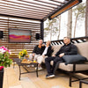Outdoor entertainment space with family relaxing under a slatted pergola, warmed by Bromic Tungsten Electric Smart-Heat heaters while enjoying drinks and a wall-mounted television.