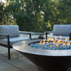 Cono Moreno 48-inch Round Copper Fire Pit set in a poolside garden during the day, surrounded by greenery and comfortable seating