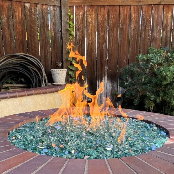 Outdoor circular brick fire pit with vibrant flames rising from colorful fire glass, set against a wooden fence in a cozy patio area