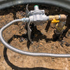 Outdoor gas line installation with a gas regulator, corrugated flexible gas hose, and a yellow valve connected to a pipe, set in a dirt and gravel area