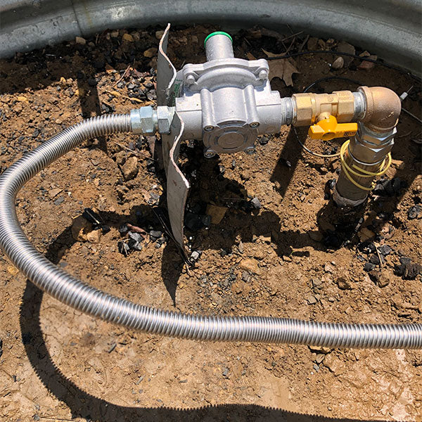 Outdoor gas line installation with a gas regulator, corrugated flexible gas hose, and a yellow valve connected to a pipe, set in a dirt and gravel area