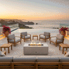 A white rectangular outdoor fire pit sits centrally on a limestone patio, surrounded by teak armchairs and sofas with grey cushions. The setting overlooks a coastal cliffside at sunset, creating a warm family gathering fire pit atmosphere. Red bougainvillea planters frame the scene, while the fire table emits a steady flame, establishing a serene backyard oasis against the ocean horizon.