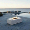 A modern fire pit in a white concrete finish rests on a bluestone patio next to a swimming pool. The rectangular fire pit is positioned in the foreground, with a lounge seating area visible in the background overlooking the ocean. Bright, clean flames demonstrate the benefits of a smokeless fire pit, enhancing the open-air ambiance of this spacious patio fire pit setup during the day.