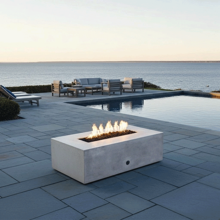 A modern fire pit in a white concrete finish rests on a bluestone patio next to a swimming pool. The rectangular fire pit is positioned in the foreground, with a lounge seating area visible in the background overlooking the ocean. Bright, clean flames demonstrate the benefits of a smokeless fire pit, enhancing the open-air ambiance of this spacious patio fire pit setup during the day.
