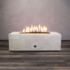 A studio presentation displays a rectangular fire pit in a smooth white finish, contrasting with a dark wood floor and textured wall. Bright orange flames flicker continuously along the linear fire pit burner, rising from a bed of black lava rocks. The visible chrome key valve on the side panel indicates the manual control of this concrete fire pit, showcasing the clean operation of a natural gas fire pit connection.