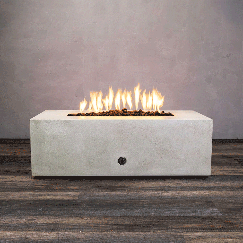 A studio presentation displays a rectangular fire pit in a smooth white finish, contrasting with a dark wood floor and textured wall. Bright orange flames flicker continuously along the linear fire pit burner, rising from a bed of black lava rocks. The visible chrome key valve on the side panel indicates the manual control of this concrete fire pit, showcasing the clean operation of a natural gas fire pit connection.