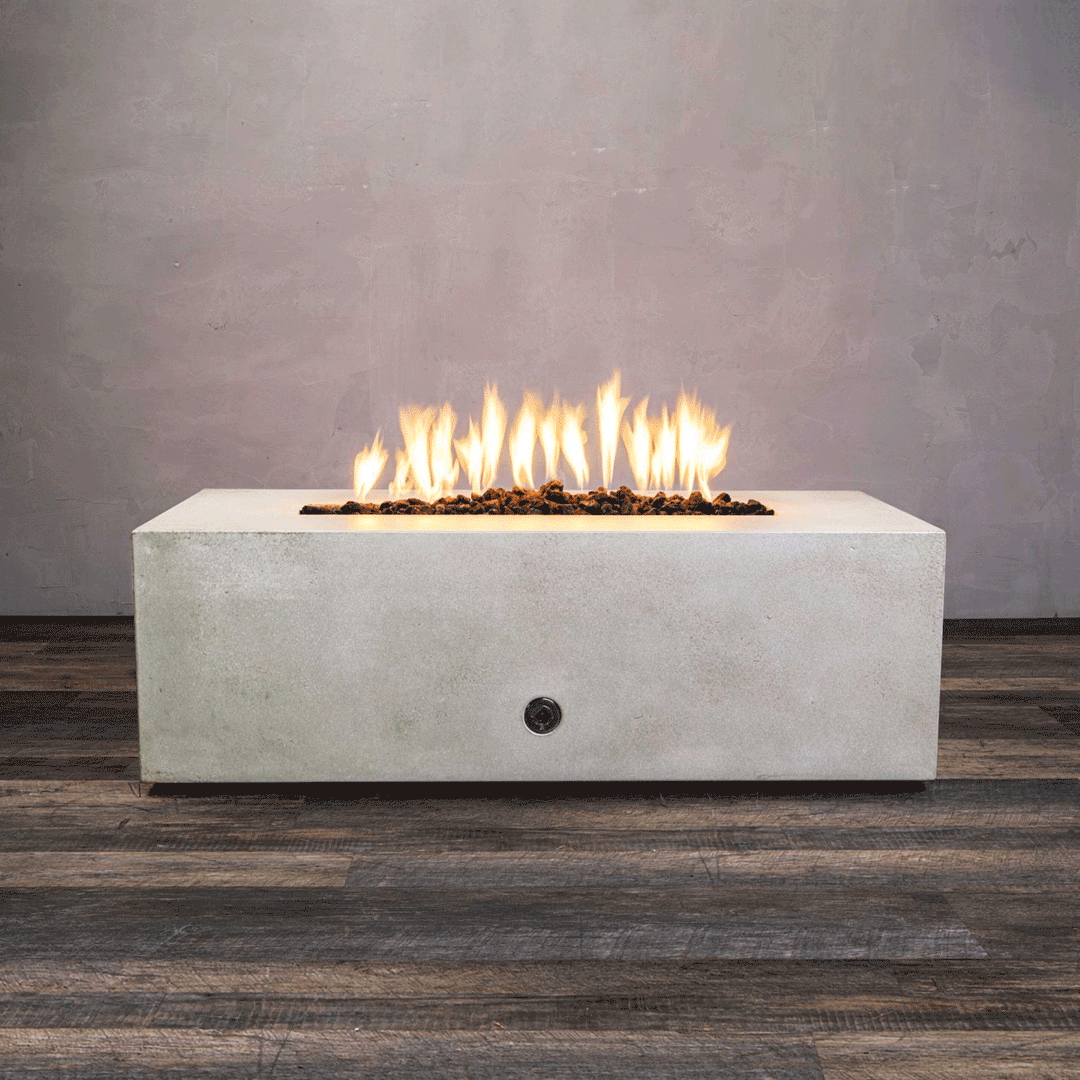A studio presentation displays a rectangular fire pit in a smooth white finish, contrasting with a dark wood floor and textured wall. Bright orange flames flicker continuously along the linear fire pit burner, rising from a bed of black lava rocks. The visible chrome key valve on the side panel indicates the manual control of this concrete fire pit, showcasing the clean operation of a natural gas fire pit connection.