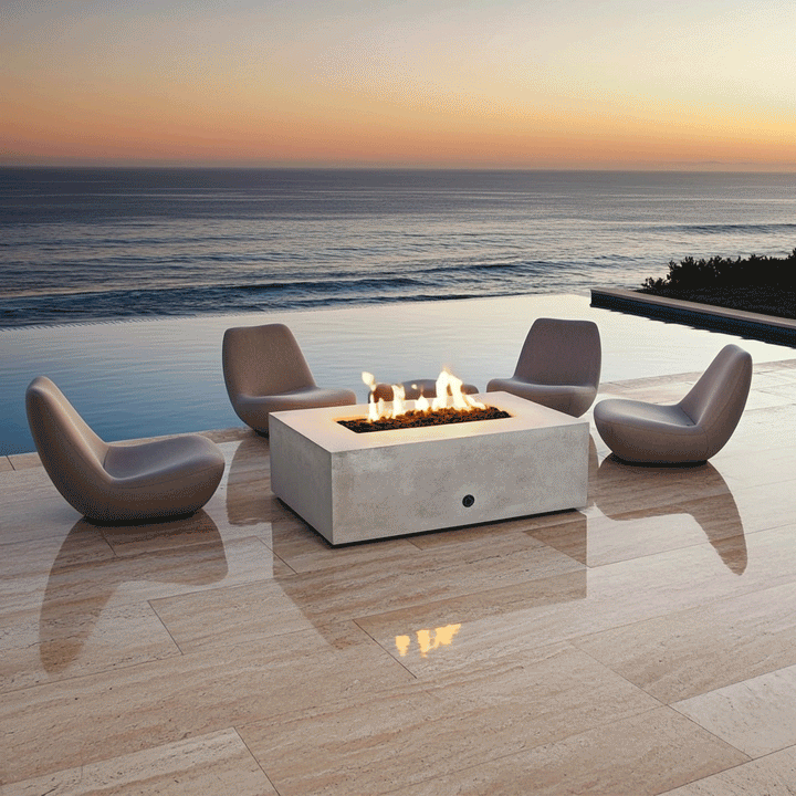 Four modern sculptural chairs surround a white luxury fire pit on a polished stone terrace overlooking the ocean at sunset. The reflection of the fire feature is visible on the glossy floor, adding depth to the scene. The steady amber flames of the propane fire pit create a focal point for outdoor entertaining, blending with the golden hues of the sky and the deep blue water.