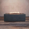 A studio presentation highlights a rectangular fire pit in a matte graphite finish, resting on a dark wood floor against a textured beige wall. Bright yellow flames dance continuously along the linear fire pit burner, rising from a bed of black lava rocks. The visible chrome key valve indicates the manual control of this natural gas fire pit, showcasing the industrial durability and sleek profile of the concrete fire pit design.