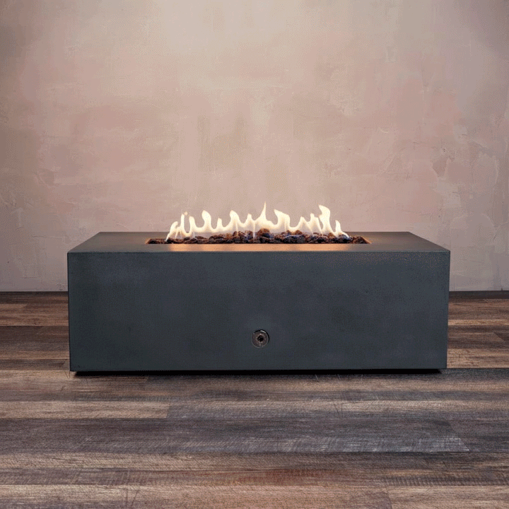 A studio presentation highlights a rectangular fire pit in a matte graphite finish, resting on a dark wood floor against a textured beige wall. Bright yellow flames dance continuously along the linear fire pit burner, rising from a bed of black lava rocks. The visible chrome key valve indicates the manual control of this natural gas fire pit, showcasing the industrial durability and sleek profile of the concrete fire pit design.