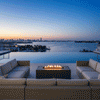 A large U-shaped sectional sofa surrounds a rectangular outdoor fire pit in a graphite finish, set against a backdrop of a harbor and city lights during blue hour. The low-profile fire table provides a warm gathering spot on the rooftop deck. Clean-burning flames illustrate the benefits of a smokeless fire pit, ensuring unobstructed views of the water while creating an inviting atmosphere for a family gathering fire pit setup.