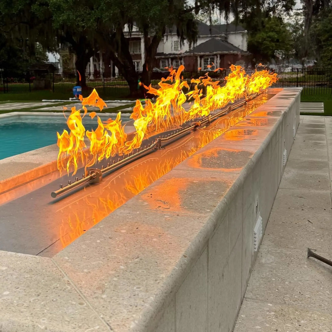 Luxurious backyard with a long, sleek linear fire feature beside a swimming pool, showcasing the brilliant flames of the HPC Fire Inspired Linear Flat Torpedo T-Burner during sunset.