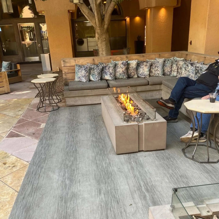 Relaxing outdoor courtyard with a modern concrete fire table and glass wind guard, featuring lively flames from the HPC Fire Inspired Linear Flat Torpedo T-Burner for a cozy seating area.