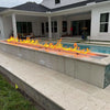 Modern outdoor living space with a long poolside fire feature illuminated by the HPC Fire Inspired Linear Flat Torpedo T-Burner, creating a warm and inviting nighttime atmosphere.