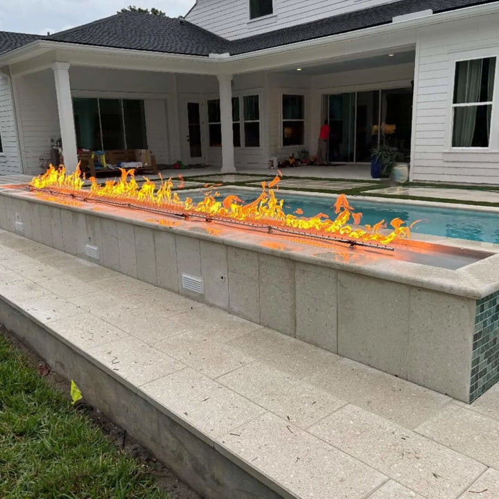 Modern outdoor living space with a long poolside fire feature illuminated by the HPC Fire Inspired Linear Flat Torpedo T-Burner, creating a warm and inviting nighttime atmosphere.