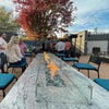 Group of people enjoying an outdoor dining space featuring a long granite fire table with glass wind guard and linear flames from the HPC Fire Inspired Linear Flat Torpedo T-Burner.