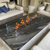 Elegant indoor-outdoor seating area with a black marble fire table featuring a glass wind guard and bright flames from the HPC Fire Inspired Linear Flat Torpedo T-Burner.