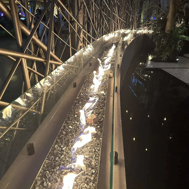 Long curved commercial fire feature with bright orange flames reflecting off black crushed glass, powered by the HPC Fire Inspired Linear Interlink Torpedo T-Burner for an elegant modern design.