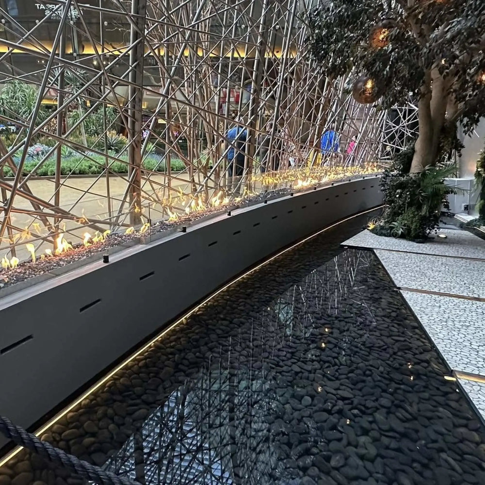 rchitectural indoor fire feature with interlinked stainless steel burners producing tall yellow flames beside a reflective water element, powered by the HPC Fire Inspired Linear Interlink Torpedo T-Burner.