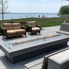 Modern outdoor patio with a rectangular linear gas fire pit featuring a glass wind guard, situated by a scenic waterfront with wicker seating, showcasing the sleek design of the HPC Fire Inspired Linear Trough Drop-In Torpedo T-Burner.