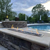Beautiful poolside setting with a stone fire feature producing bright linear flames, illustrating the performance and elegance of the HPC Fire Inspired Linear Drop-In Torpedo T-Burner for backyard retreats.
