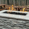 Rectangular stone fire pit with realistic lava rocks and vivid flames, showcasing the power and beauty of the HPC Fire Inspired Linear Trough Drop-In Torpedo T-Burner on a patio with wooden picnic benches nearby.
