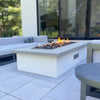Modern outdoor living area featuring the Hearth Products Controls (HPC) Fire Inspired Rectangle Drop-In Burner set into a sleek rectangular fire pit. The fire pit has a white stucco base with a smooth gray concrete top, and is filled with black lava rocks producing a vibrant, natural flame—perfectly complementing the contemporary patio seating and creating a cozy gathering space.