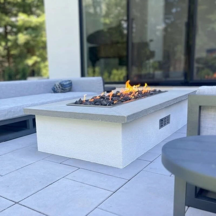 Modern outdoor living area featuring the Hearth Products Controls (HPC) Fire Inspired Rectangle Drop-In Burner set into a sleek rectangular fire pit. The fire pit has a white stucco base with a smooth gray concrete top, and is filled with black lava rocks producing a vibrant, natural flame—perfectly complementing the contemporary patio seating and creating a cozy gathering space.