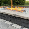 The Hearth Products Controls (HPC) Fire Inspired Rectangle Drop-In Burner is beautifully installed in a custom-built outdoor fire feature. Surrounded by natural stone tile and a protective glass wind guard, the burner is filled with smooth lava rocks and produces vibrant flames—perfectly blending with the multi-toned stonework and creating a cozy, stylish centerpiece for modern patio settings.