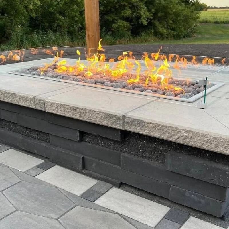 The Hearth Products Controls (HPC) Fire Inspired Rectangle Drop-In Burner is beautifully installed in a custom-built outdoor fire feature. Surrounded by natural stone tile and a protective glass wind guard, the burner is filled with smooth lava rocks and produces vibrant flames—perfectly blending with the multi-toned stonework and creating a cozy, stylish centerpiece for modern patio settings.