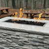 Outdoor fire feature showcasing the Hearth Products Controls (HPC) Fire Inspired Rectangle Drop-In Burner filled with black lava rocks, set in a polished stone top with a textured gray stacked stone base. Surrounded by picnic benches and forest scenery, this modern yet rustic setup adds cozy ambiance and warmth to outdoor gatherings.