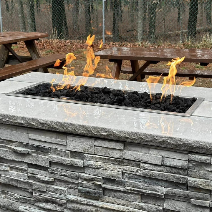 Outdoor fire feature showcasing the Hearth Products Controls (HPC) Fire Inspired Rectangle Drop-In Burner filled with black lava rocks, set in a polished stone top with a textured gray stacked stone base. Surrounded by picnic benches and forest scenery, this modern yet rustic setup adds cozy ambiance and warmth to outdoor gatherings.