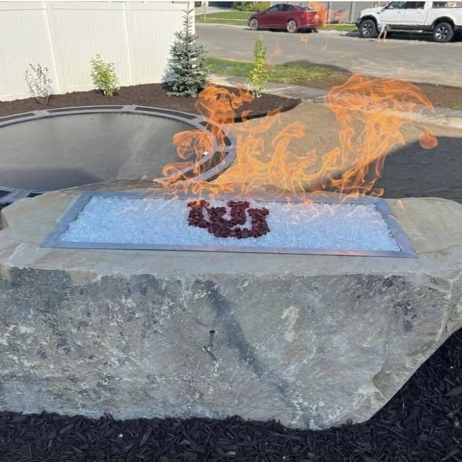 The Hearth Products Controls (HPC) Fire Inspired Rectangle Drop-In Burner is installed in a custom outdoor fire feature carved from a large natural boulder. The burner is filled with clear fire glass and features a striking red "U" logo in the center, creating a dramatic flame display—perfect for showcasing school spirit in a rustic yet modern backyard setting.