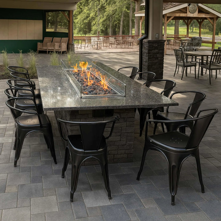 Rectangular granite fire table featuring an HPC Fire Inspired stainless steel burner with vibrant flames, surrounded by black metal chairs in a covered outdoor patio dining space.