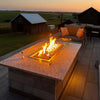 Elegant outdoor patio setup showcasing an HPC Fire Inspired rectangular drop-in burner set in a granite fire table, glowing beautifully at sunset beside a cozy seating area and scenic barn view.