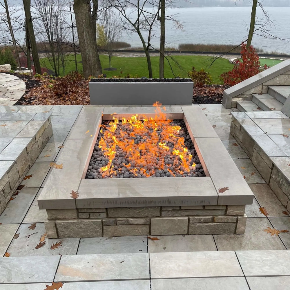 Modern outdoor patio with a square stone fire pit featuring an HPC Fire Inspired gas burner producing bright, natural flames across lava rocks. The setup overlooks a scenic lake and landscaped yard, creating a warm and inviting gathering space perfect for cool evenings.