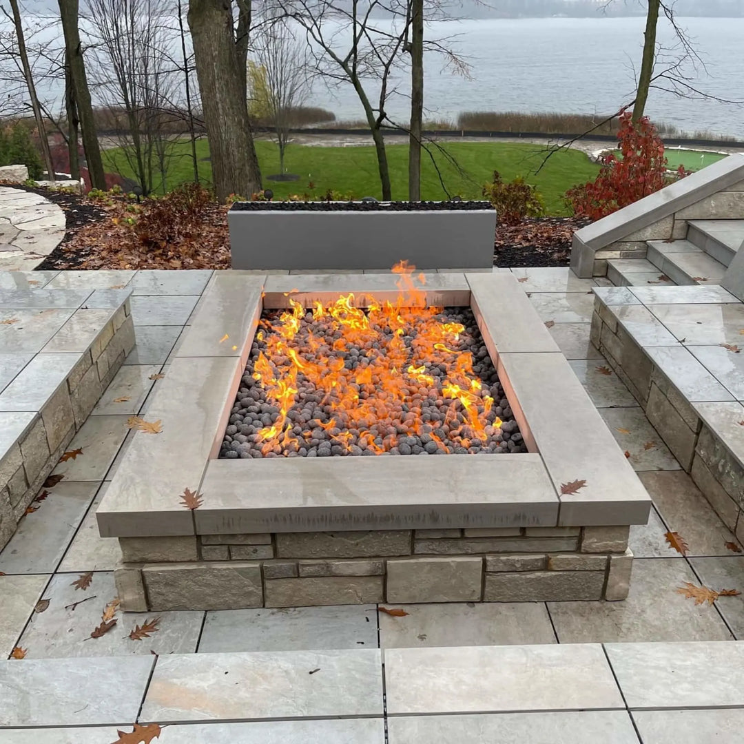 Modern outdoor patio with a square stone fire pit featuring an HPC Fire Inspired gas burner producing bright, natural flames across lava rocks. The setup overlooks a scenic lake and landscaped yard, creating a warm and inviting gathering space perfect for cool evenings.