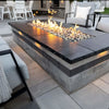 Contemporary outdoor patio featuring a sleek rectangular stone fire pit with bright flames over reflective fire glass, creating a warm and modern atmosphere for evening relaxation.
