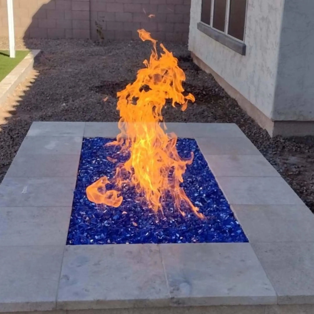 Rectangular backyard fire pit with bold orange flames rising from cobalt blue fire glass, offering a striking contrast against light stone tile for a stylish outdoor centerpiece.