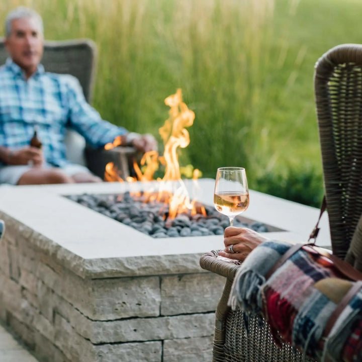 Couple relaxing by a stone fire pit with glowing flames over gray lava rocks, enjoying a peaceful evening outdoors with drinks and warm ambiance.