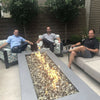 Three men sitting comfortably around a rectangular fire table with clear fire glass media, enjoying casual conversation and warmth from the HPC Fire Inspired burner.