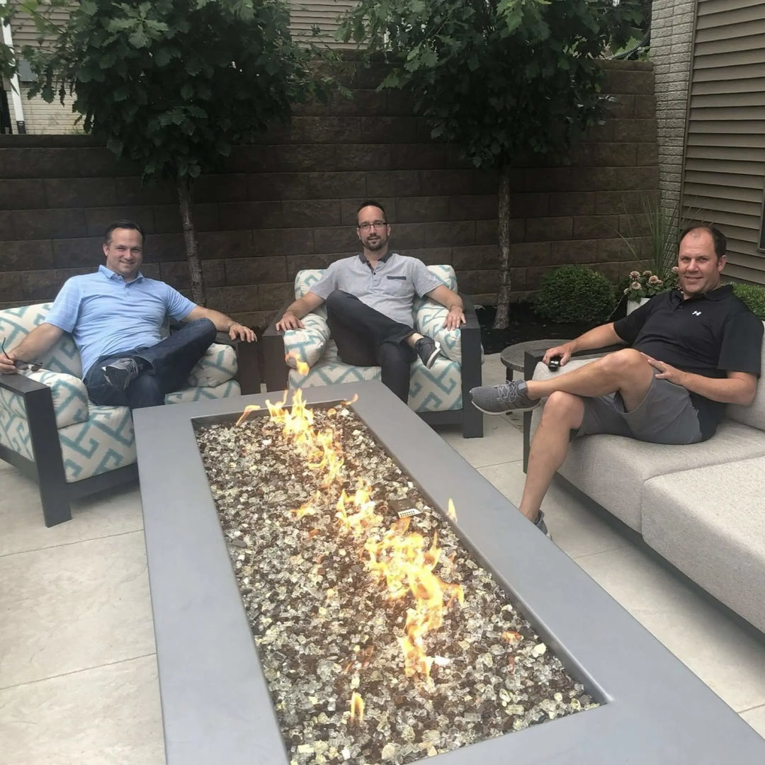 Three men sitting comfortably around a rectangular fire table with clear fire glass media, enjoying casual conversation and warmth from the HPC Fire Inspired burner.