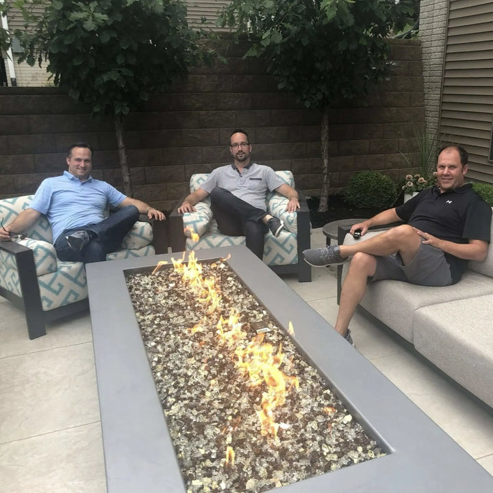 Three men sitting comfortably around a rectangular fire table with clear fire glass media, enjoying casual conversation and warmth from the HPC Fire Inspired burner.