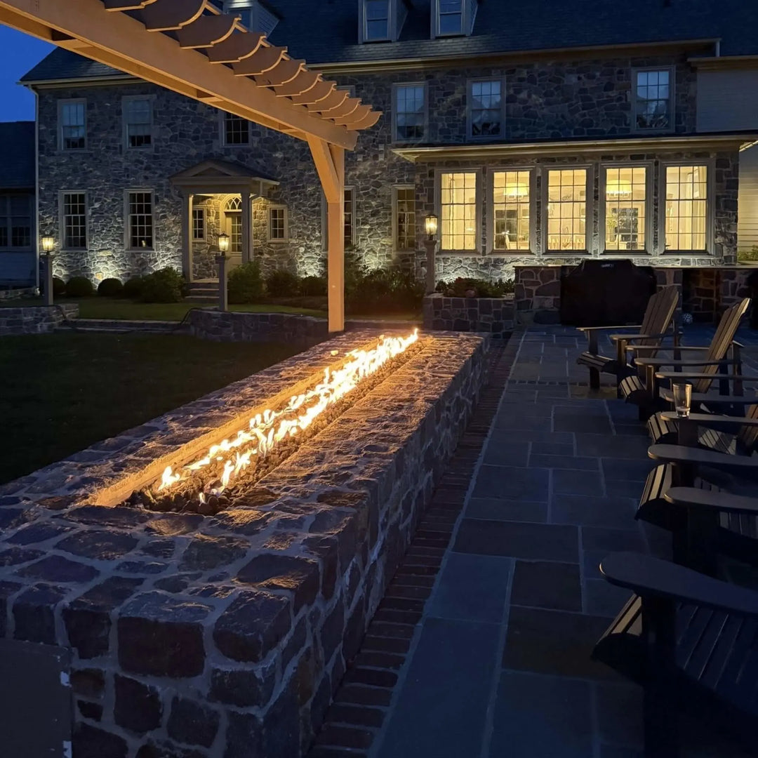 Long stone fire feature with bright linear flames casting a warm glow on a traditional patio, complementing the elegant stone exterior of a large home at dusk.