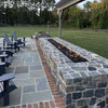 Daytime view of a rustic stone patio with a rectangular fire pit framed by dark lava rock, surrounded by Adirondack chairs and scenic countryside greenery.
